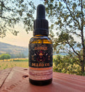 A 1 fl oz amber glass dropper bottle of Gorge Beard Co. Bergamot Woods beard oil, set on a rusty metal surface with a blurred outdoor landscape of trees and hills in the background.